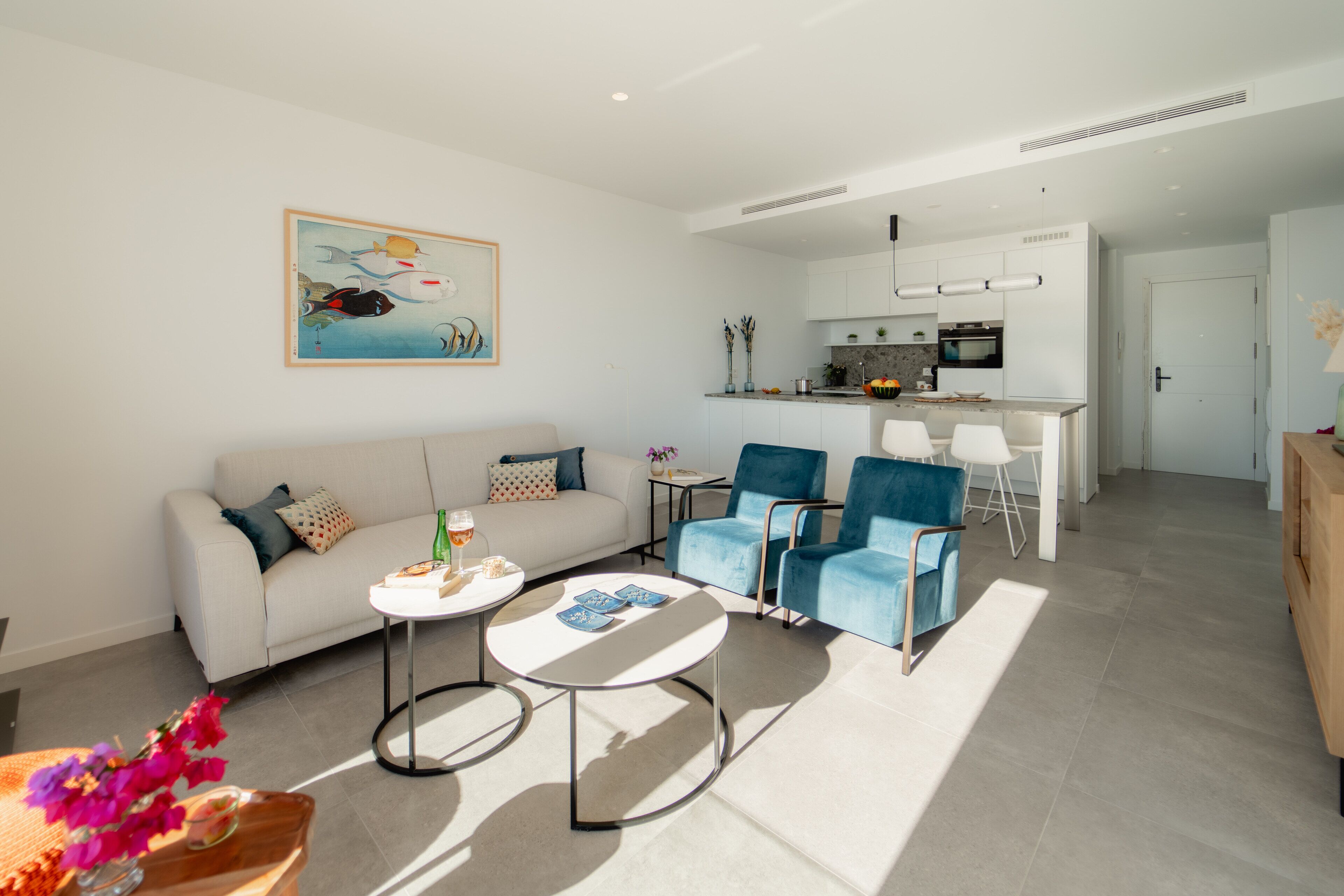 Una sala de estar moderna con sofá, mesa de centro y sillas azules. Se observa una zona de cocina con armarios blancos y una mesa de comedor.