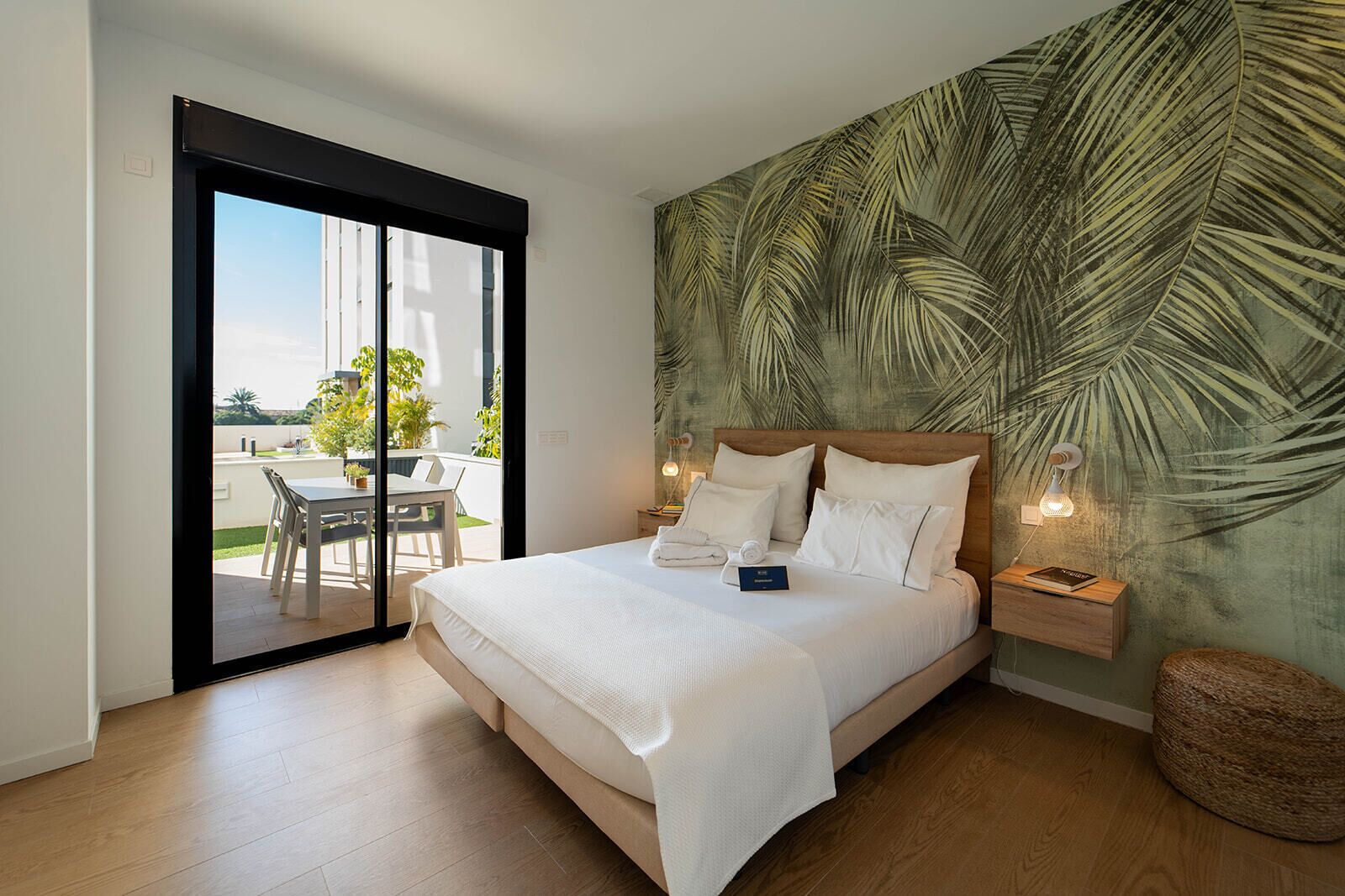 A hotel room with a large bed, a wooden headboard, a sliding glass door to a balcony, a tropical leaf-patterned wall, and a small bedside table with a lamp.