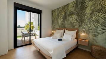 A hotel room with a large bed, a wooden headboard, a sliding glass door to a balcony, a tropical leaf-patterned wall, and a small bedside table with a lamp.