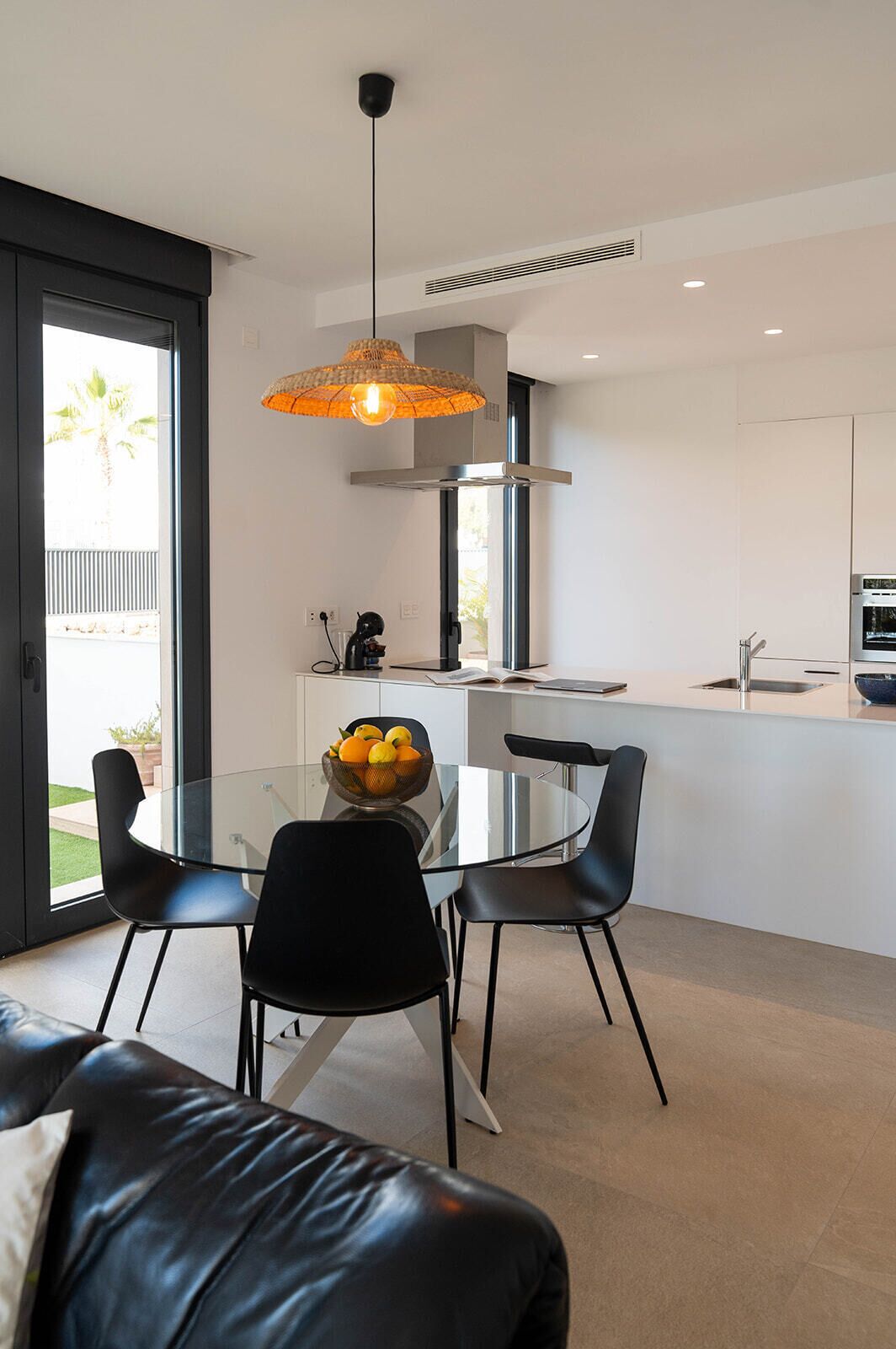A modern kitchen with a glass dining table, black chairs, and a fruit bowl.