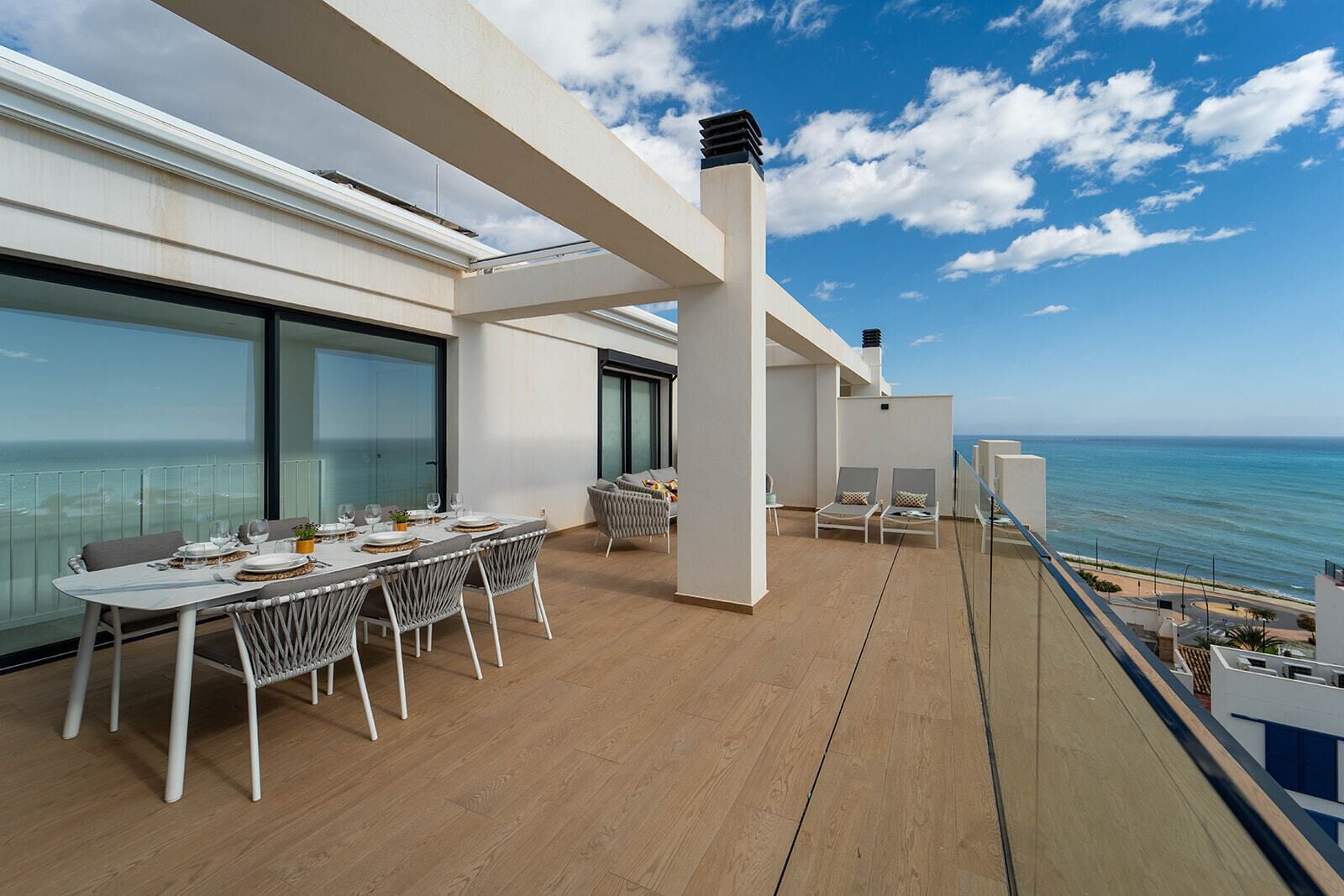 Un balcón con una mesa de comedor preparada para cuatro personas, dos sillones de descanso y vistas al mar.
