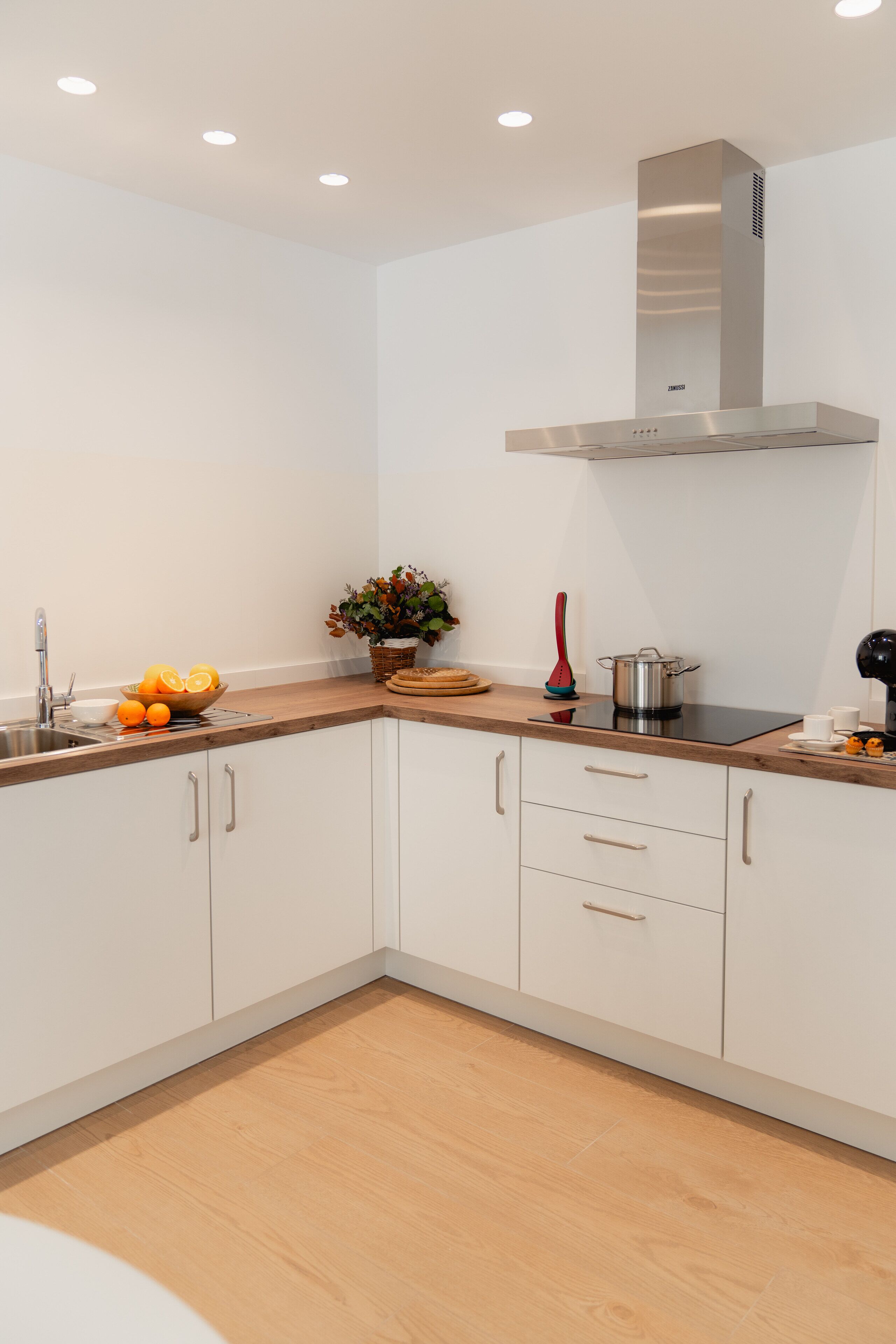 A modern kitchen with white cabinets, a stainless steel range hood, a sink, and a countertop with fruits and a plant.