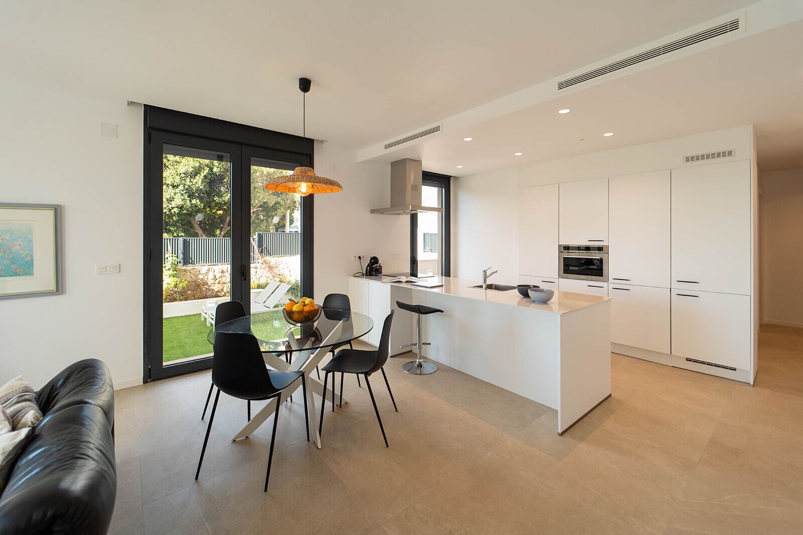 Una cocina moderna con una isla central, un comedor con mesa de vidrio, sillas negras y una lámpara colgante.