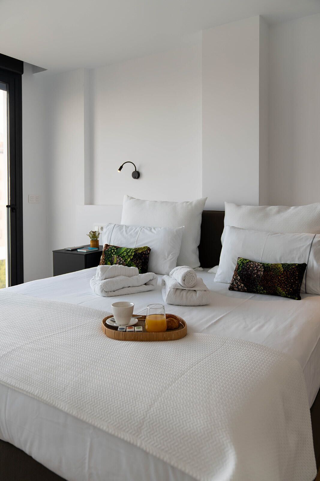 Een netjes opgemaakt bed met witte lakens, een dienblad met ontbijtartikelen en een wandlamp.