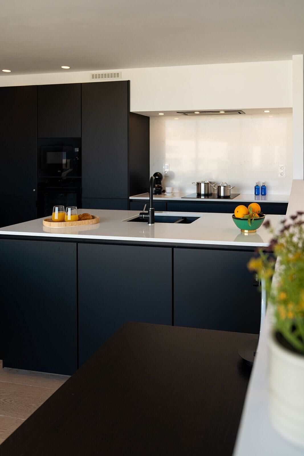 Una cocina moderna con armarios oscuros, una encimera blanca y una isla central. Hay un cuenco con naranjas en la isla y una máquina de café en la encimera.