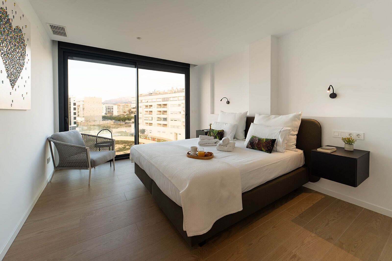 A modern bedroom with a large bed, a chair, a nightstand, and a view of buildings through a large window.