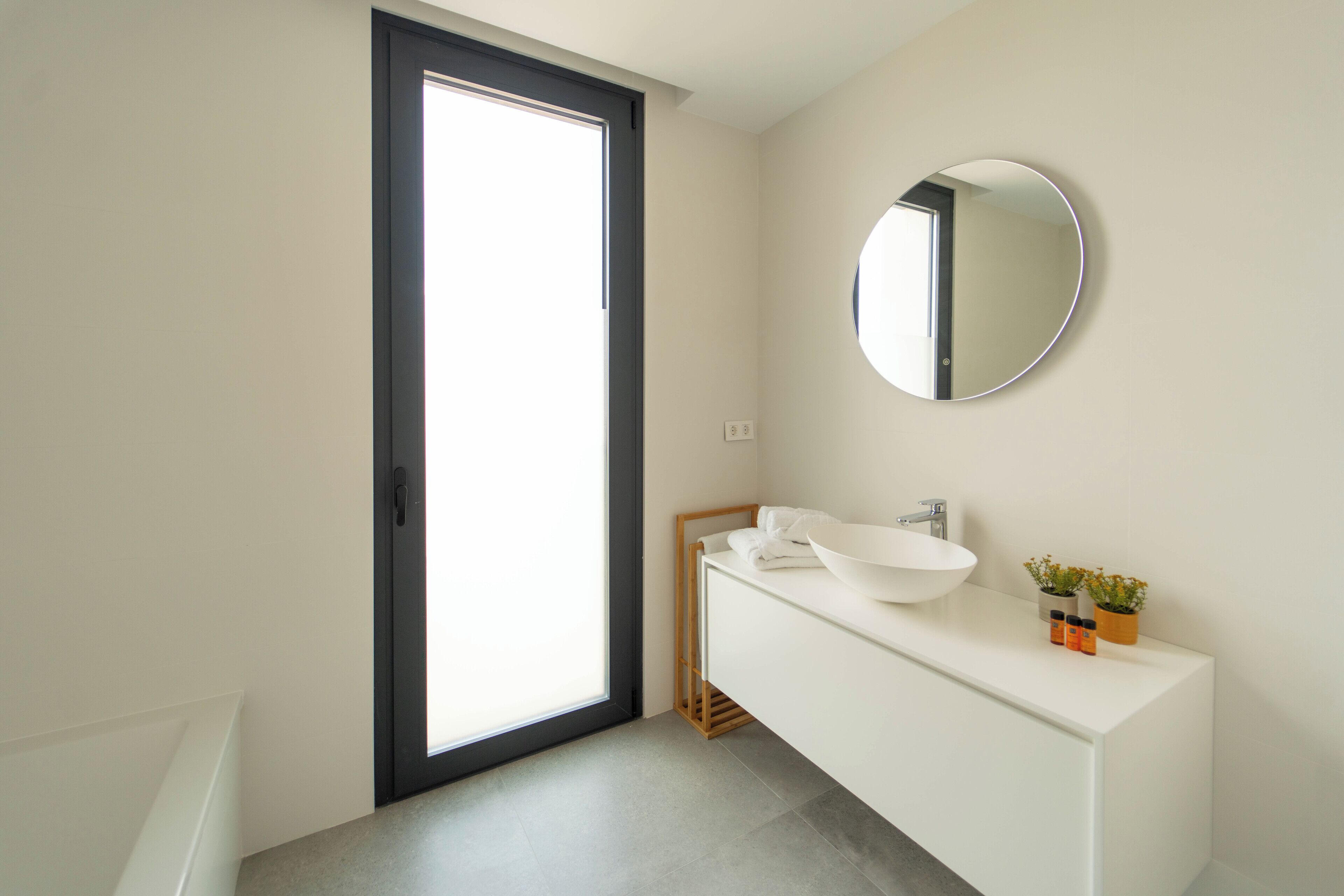 A modern bathroom with a large window, a round mirror, a white sink, and a small plant on the vanity.