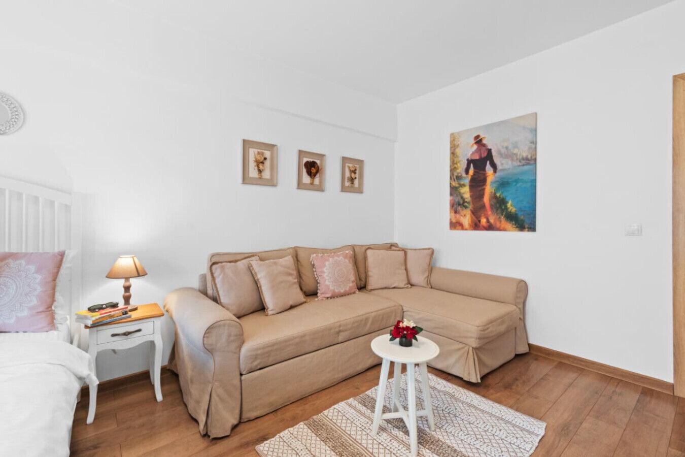 Ett sovrum med en beige soffa, ett sängbord med en lampa, en liten pall och en inramad tavla på väggen.