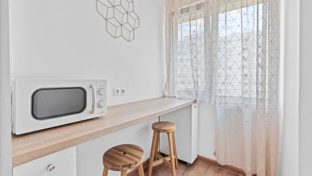 A kitchenette with a microwave, wooden stools, a geometric wall decoration, and a window with sheer curtains. - Sudului 507 by MRG Apartments (București)