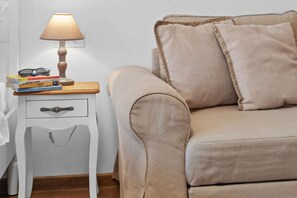 A bedroom with a beige sofa, a bedside table with a lamp, and a stack of books.