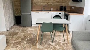 A compact kitchen with a stone wall, a refrigerator, a washing machine, and a dining area with chairs. - Appartement Lumineux en Rdc, Pour Séjour Touristique ou Professionnel (Bourgoin-Jallieu)