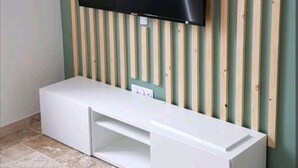 A room with a flat-screen TV mounted on a wooden slat wall, a white entertainment unit below, and tiled flooring. - Appartement Lumineux en Rdc, Pour Séjour Touristique ou Professionnel (Bourgoin-Jallieu)