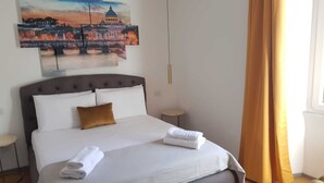 A neatly made bed with white linens and a brown pillow, a bedside table, and a painting of a cityscape on the wall.