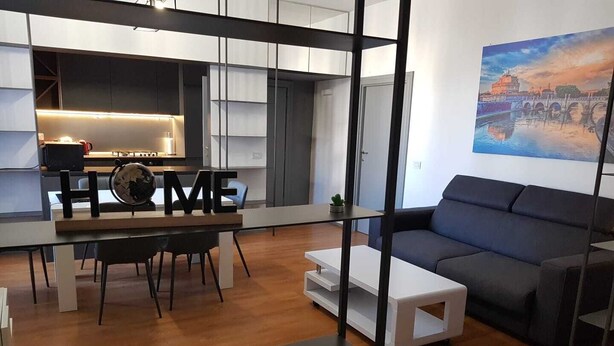 A modern living room with a dark sofa, a coffee table, and a dining area with a table and chairs. There is a kitchen in the background with a TV and a small plant on the counter.