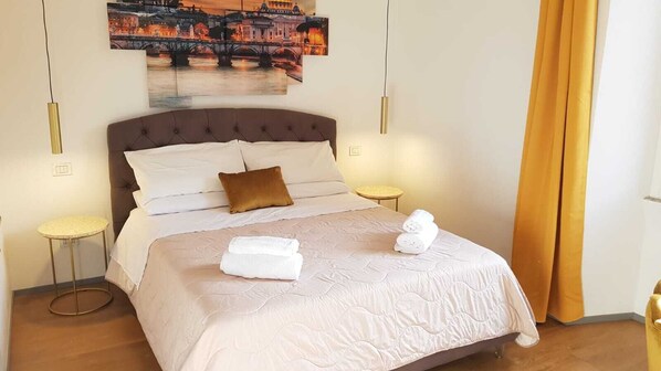 Un lit bien fait, avec du linge de lit blanc et deux serviettes, une tête de lit marron et, au-dessus, un tableau représentant un paysage urbain.