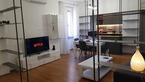 Un salon moderne doté d’un téléviseur à écran plat affichant le logo « NETFLIX », d’un coin repas avec une table et des chaises, et d’une cuisine avec un plan de travail et des placards.