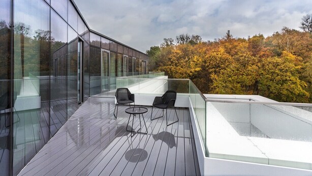 Een modern terras buiten met glazen balustrades, twee zwarte stoelen en een houten vloer.