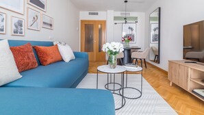 A modern living room with a blue sofa, a small round table with a vase of flowers, a dining area with a table and chairs, and a TV on a wooden cabinet. - López de Hoyos II (Madrid)
