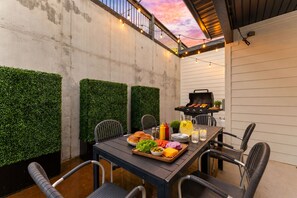 Een terras buiten, met een tafel gedekt voor een maaltijd, omringd door stoelen, een barbecue en sfeerverlichting.