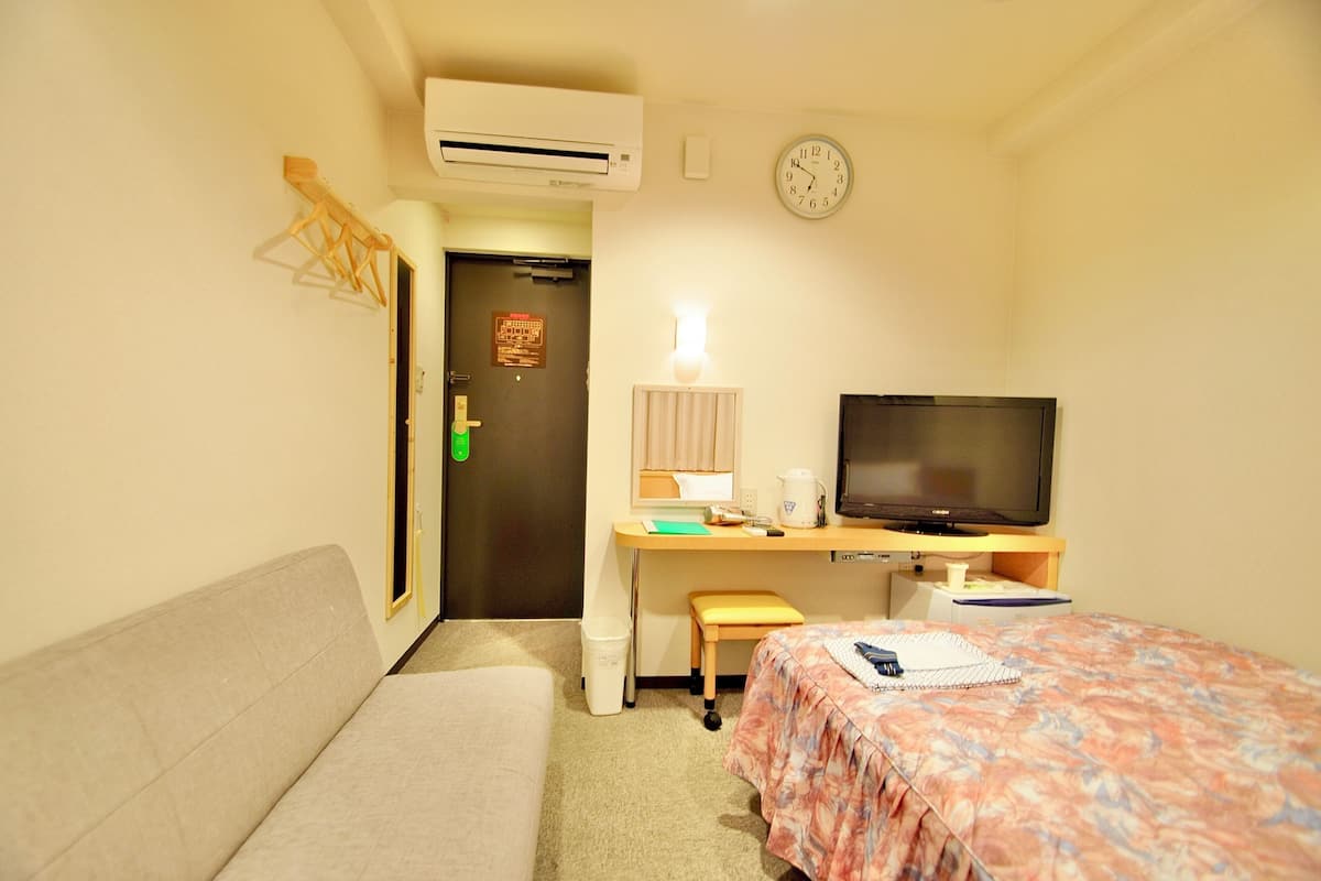 A hotel room with a bed, a desk with a television, a sofa, and a wall clock.