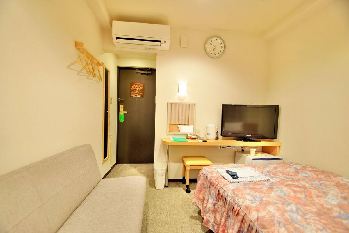 Ein Hotelzimmer mit einem Bett, einem Schreibtisch mit Fernseher, einer Couch und einer Wanduhr.