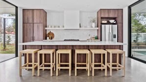 A modern kitchen with a central island, wooden stools, and a stainless steel refrigerator. - Maple Suite, Berry - by Linnaeus Collection (Berry)