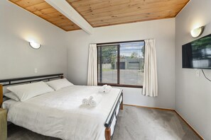 A bedroom with a bed, a window with curtains, a wall-mounted TV, and a ceiling with wooden beams.