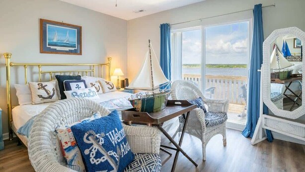 A bedroom with a bed, a chair, a table, a ceiling fan, a window with curtains, and a framed picture on the wall. - Jolly Roger (North Topsail Beach)