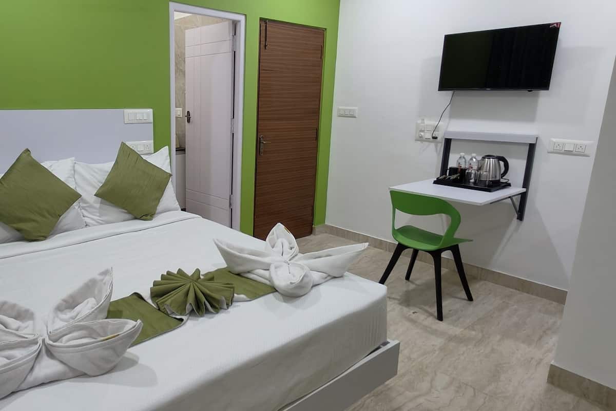 Una cama bien hecha con almohadas blancas y verdes, una silla verde, un escritorio pequeño con un hervidor y un televisor en la pared.