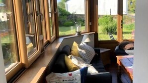 A cozy room with a sofa and a table, large windows offering a view of the garden and a white building outside.