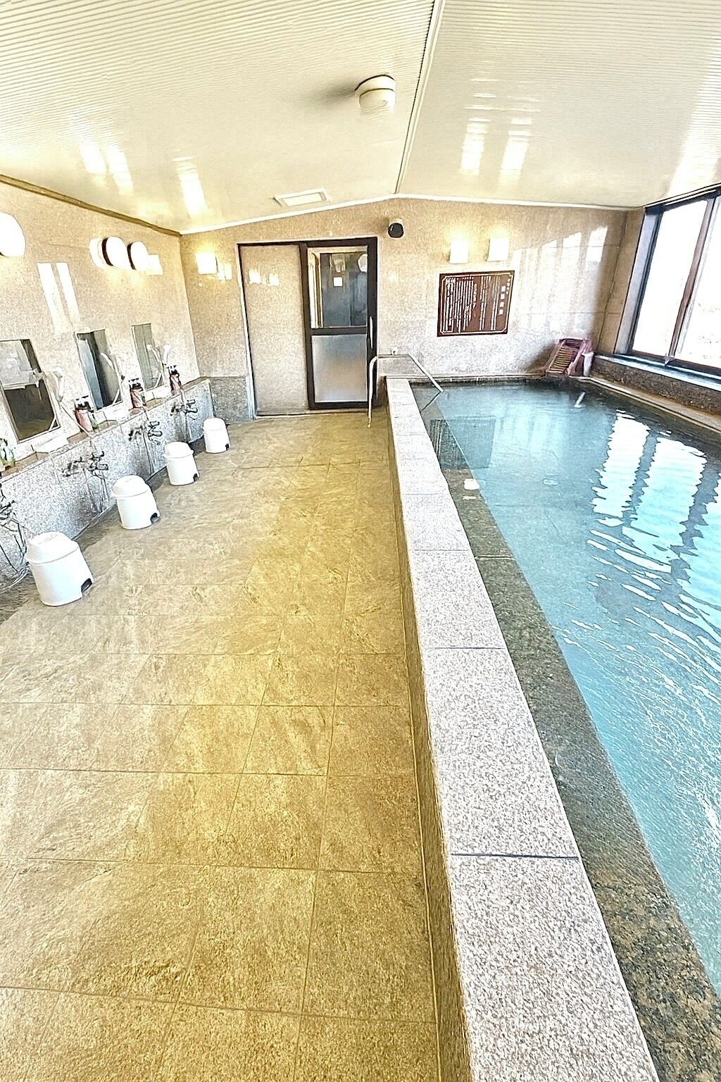 Uima-allas, jonka pohja on laatoitettu ja jossa on lasinen kaide. Seinää pitkin on useita pesualtaita, joissa on peilit ja hanat.