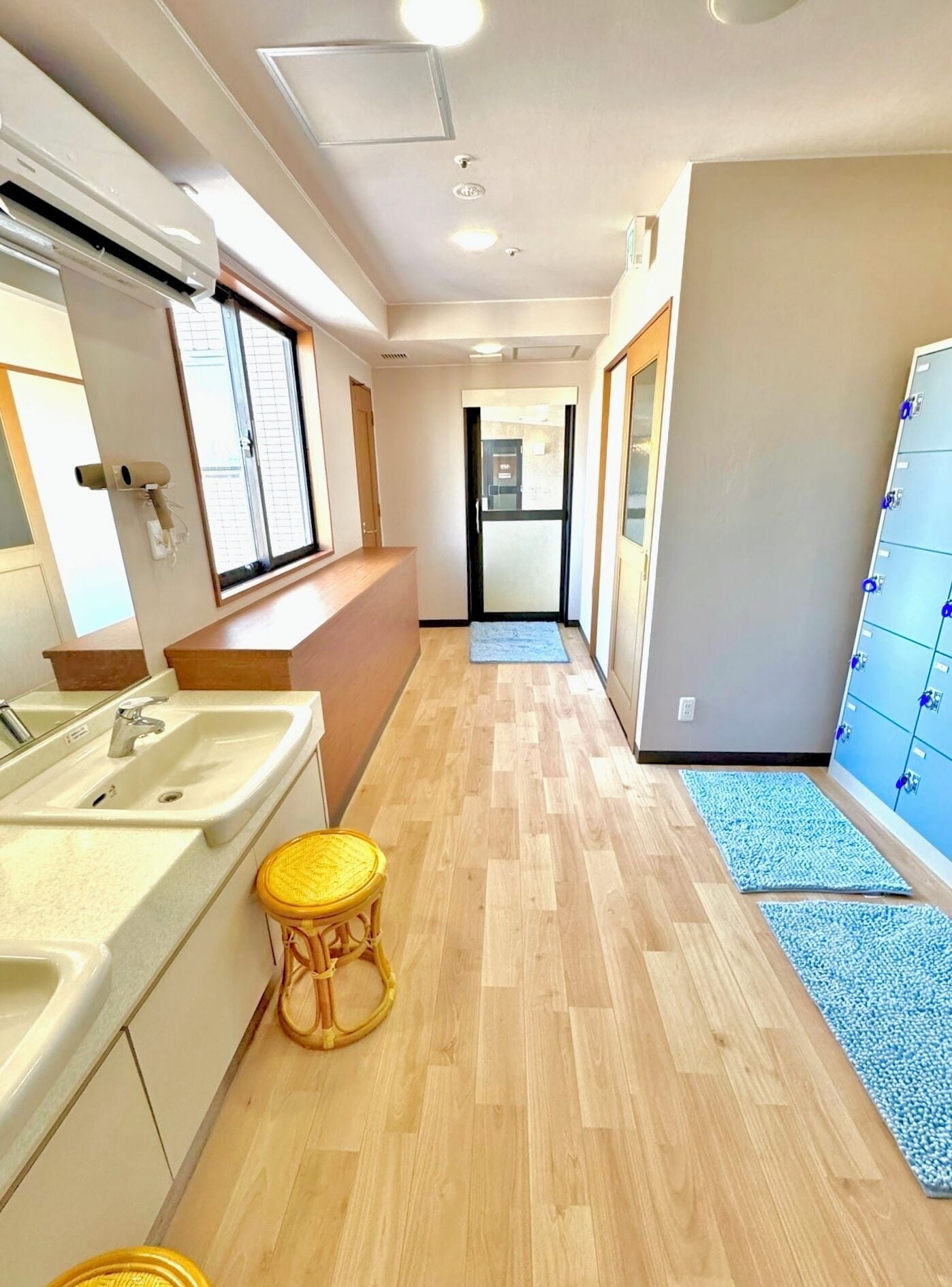 A hotel hallway with a wooden floor, a bathroom with a sink and mirror, and a blue mat leading to a door.
