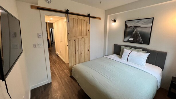 A bedroom with a bed, a wooden door, a framed picture, and a hallway leading to another room. - Dt 1Bed 1bath Cozy Suite 8 Min Walk To Arena (Edmonton)