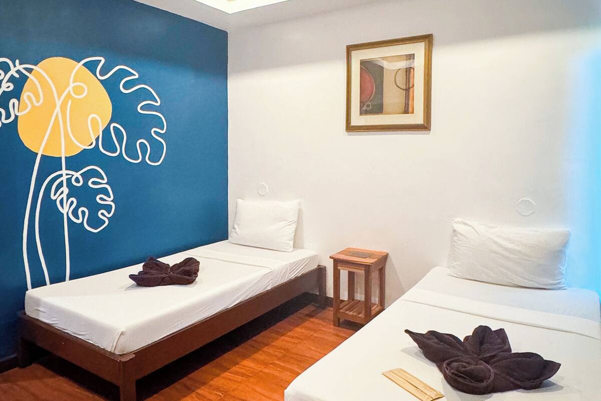 Ein Hotelzimmer mit zwei Einzelbetten, Holzboden, einer blauen Wand mit floralem Dekor, einem kleinen Nachttisch aus Holz und einem gerahmten Bild an der Wand.