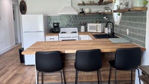 Ett modernt kök med en bänkskiva i trä, två svarta barstolar, ett vitt kylskåp, en spis och en mikrovågsugn.