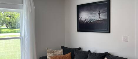 A modern living room with a grey sofa, two patterned cushions, a framed picture of a lighthouse, and a window with white curtains.