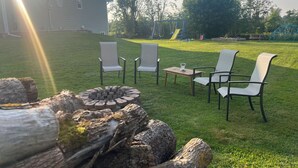 En trädgård med en eldstad, tre stolar och ett hus i bakgrunden.