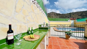 Eine Terrasse mit Pool, einer Steinsäule, einem Tisch mit einer Weinflasche, Gläsern und Snacks.