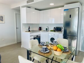 Cucina moderna con mobili bianchi, frigorifero in acciaio inossidabile, microonde e lavatrice. Si nota un tavolo da pranzo in vetro con sedie, un cesto di frutta e una bottiglia di vino.