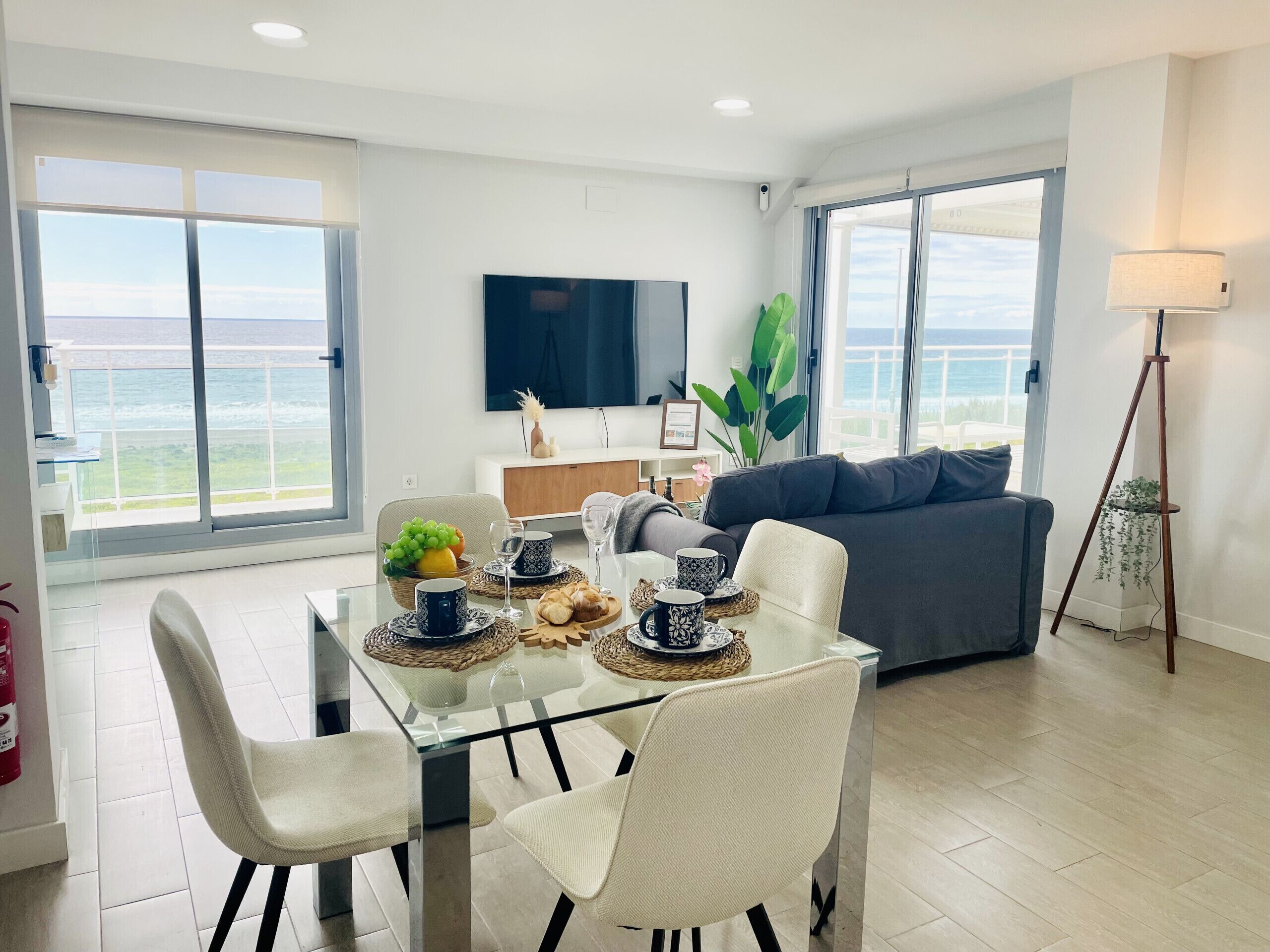 Una sala de estar moderna con una mesa de comedor de cristal, sillas blancas, un sofá gris y un amplio ventanal con vista al océano.
