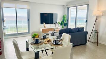Una sala de estar moderna con una mesa de comedor de cristal, sillas blancas, un sofá gris y un amplio ventanal con vista al océano.