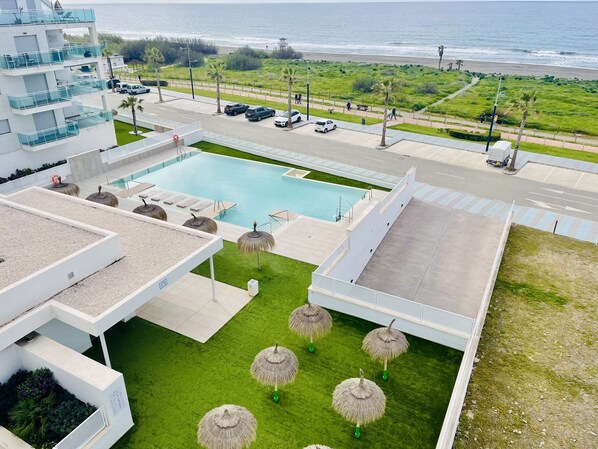 A pool area with a view of the ocean, surrounded by a modern building with balconies and a parking area.