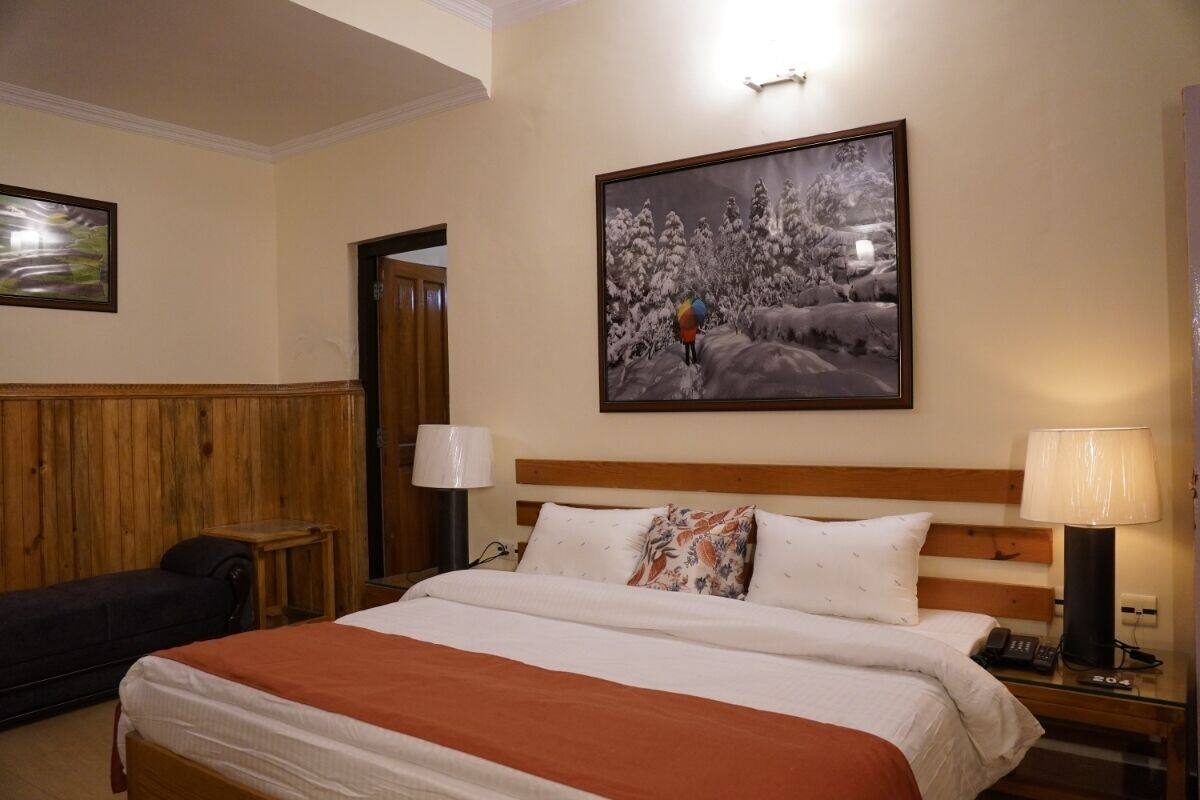 A hotel room with a large bed, a wooden headboard, a nightstand with a lamp, and a framed picture of a snowy landscape on the wall.