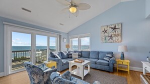 A spacious living room with a large window overlooking the ocean, a ceiling fan, and a view of the beach. - A Southern Charm - 5BR Oceanfront House in Surf City Sleeps 12 (Surf City)