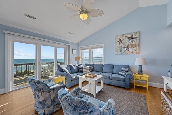 A spacious living room with a large window overlooking the ocean, a ceiling fan, and a view of the beach.