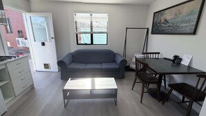 A living room with a sofa, a dining table with chairs, a coffee table, a window with blinds, and a painting on the wall. - Private Studio | Convenient Stay Near Hobart (Claremont)