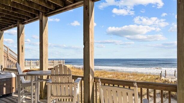 Una terraza de madera con dos sillas Adirondack y una parrilla, con vista a una playa que tiene un paseo de madera y vista al océano.