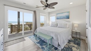 Une chambre avec un grand lit, un ventilateur de plafond, une vue sur la plage et un tableau représentant l’océan.