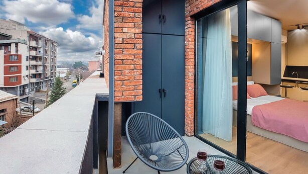 Ein modernes Hotelzimmer mit Ziegelaußenwand, einer großen Schiebeglasdoor, einem Bett mit rosa Bettwäsche, einem kleinen Tisch und einem Stuhl sowie Blick auf die Stadtlandschaft.