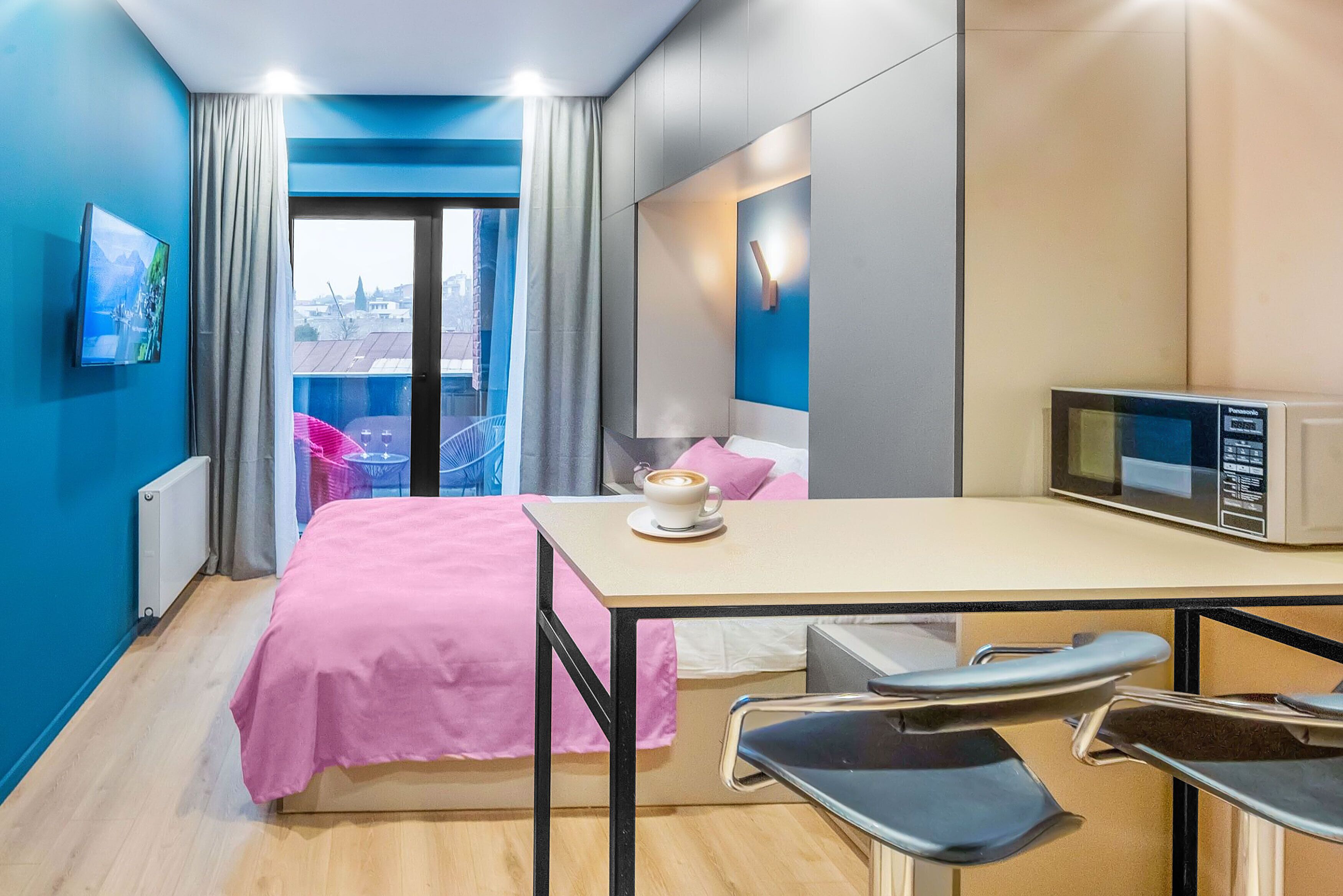 A modern hotel room with a bed, a desk, a chair, a microwave, and a view of the city.